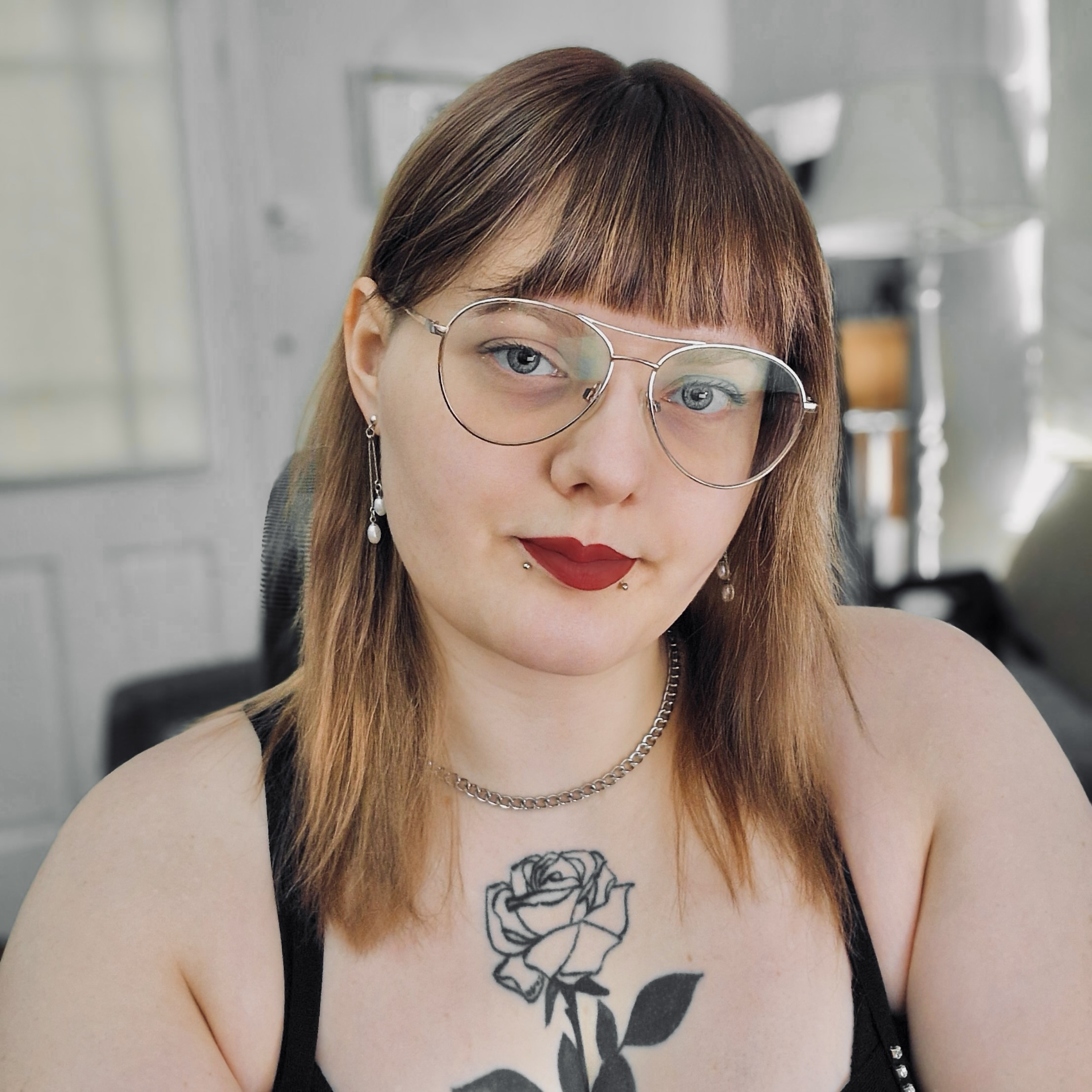 headshot of Ennis Welbourne, a white person wearing glasses and lipstick with a black line rose tattooed on the chest