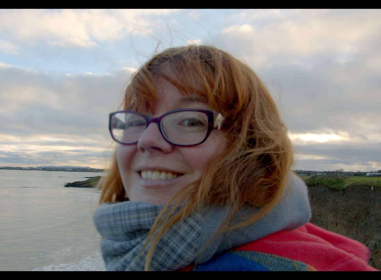 headshot of Órla Price, a ginger person with glasses and a scarf, with a coastal line in the background